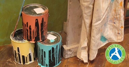 Three paint cans with one stacked on top of the other two and paint falling down the side next to a white drop cloth covered in paint
